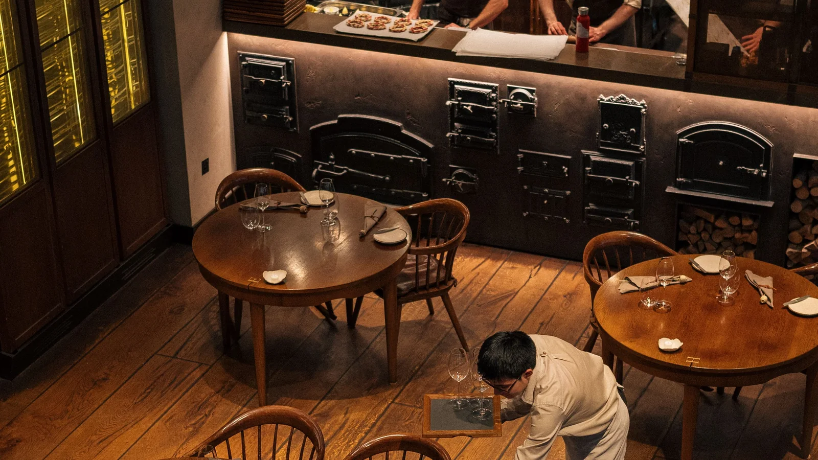 Koks aan het werk in een open keuken terwijl een kelner de tafels dekt in een warm verlicht restaurant