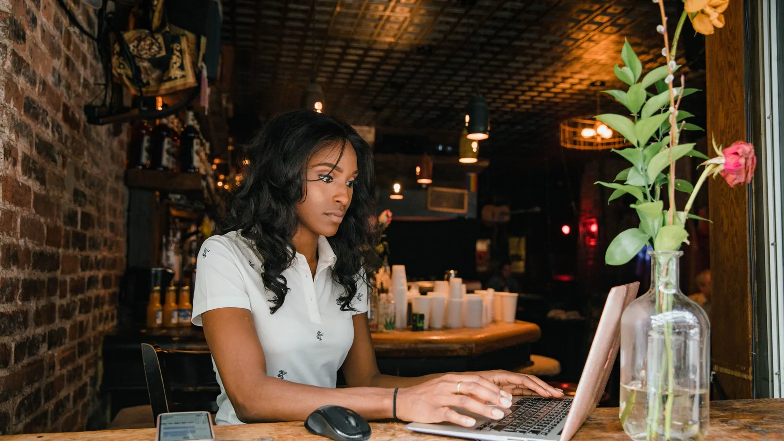 Jonge vrouw werkt op haar laptop aan de houten toog van een café met bakstenen muren