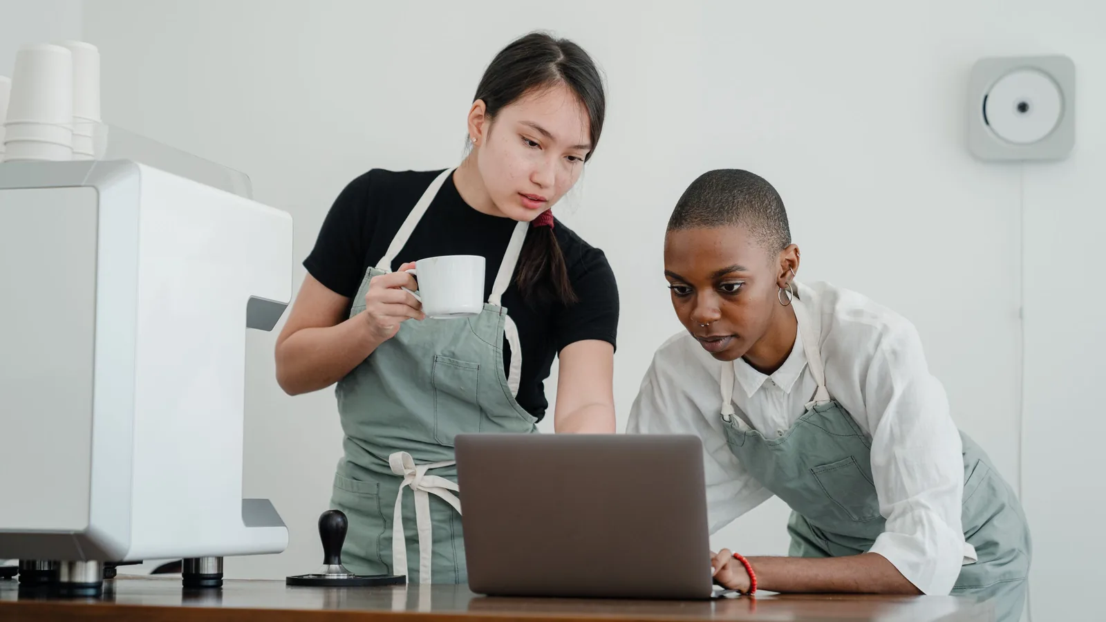 Twee medewerkers in schort bekijken samen een laptop aan de toog naast de koffiemachine