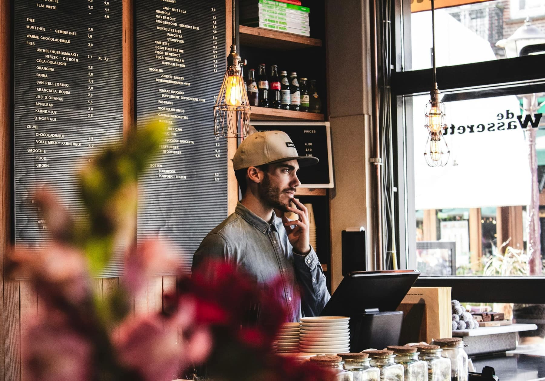 Een uitbater achter de kassa van zijn brasserie, met het menubord achter hem